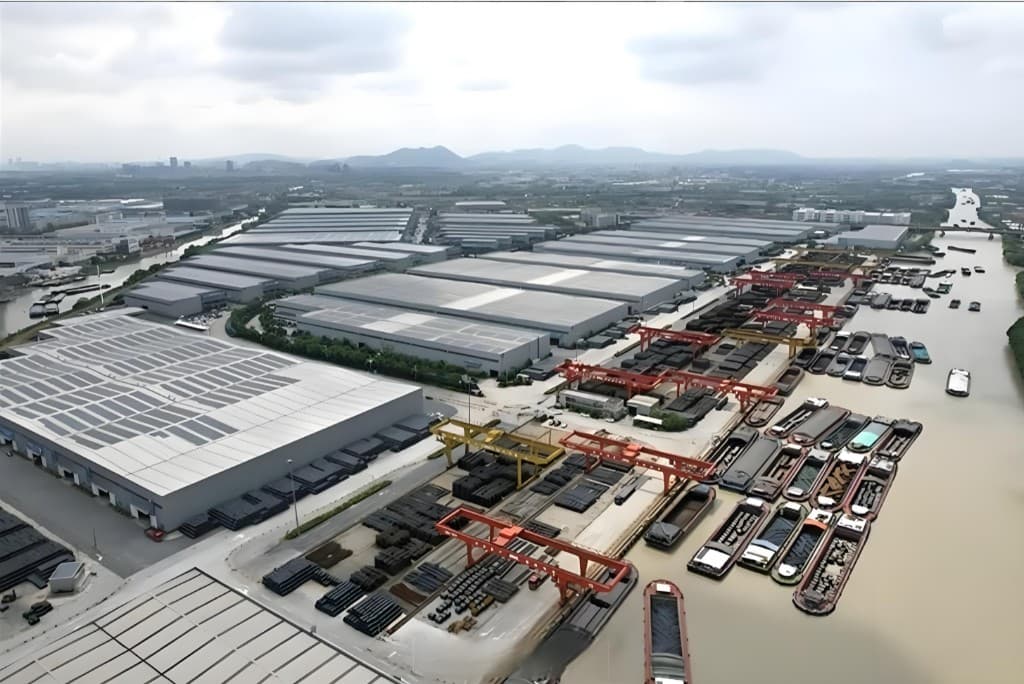 Aerial view of the JURUN plant — vast covered warehouses, gantry-crane storage yards, and a canal lined with cargo barges loading steel pipe